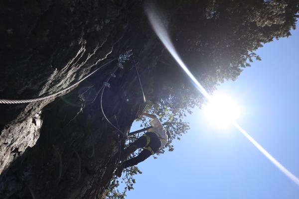 A person is seen actively climbing a rugged rock face, secured by a harness and cables, under a bright blue sky